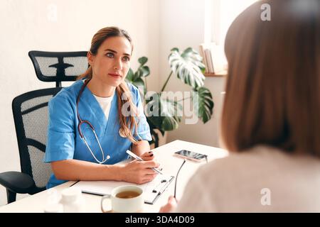 Medico donna in uniforme medica con stetoscopio seduto a tavolo con laptop prendendo appunti e parlando con il paziente nello studio medico in ospedale. Ciao Foto Stock
