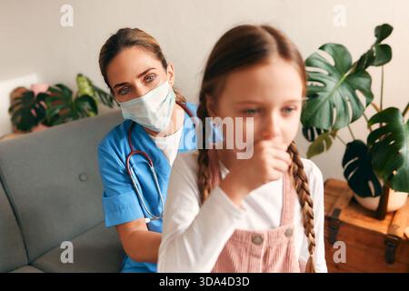 Medico di famiglia a casa in una maschera protettiva medica ascoltare i polmoni di un bambino seduto su un divano nel soggiorno. Esame del paziente, guarigione Foto Stock
