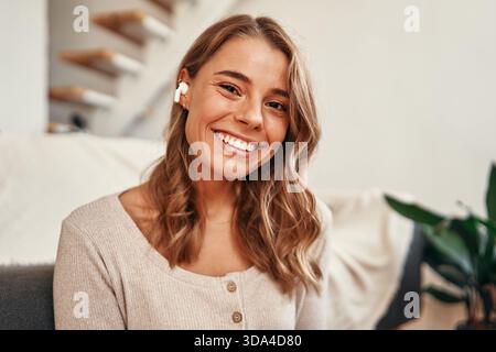 Giovane donna che ascolta musica o lezione audio con le cuffie mentre è seduta sul pavimento vicino al divano nel salotto di casa. Foto Stock