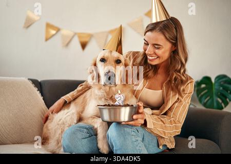 Giovane donna felice che tiene la torta di compleanno, festeggia il compleanno del suo cane da compagnia, abbraccia e si diverte con il suo cucciolo seduto in salotto a h Foto Stock