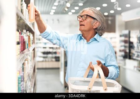Uomo anziano con capelli e occhiali grigi in piedi in una farmacia, tenendo un cestino bianco e raggiungendo una bottiglia dallo scaffale pieno di va Foto Stock