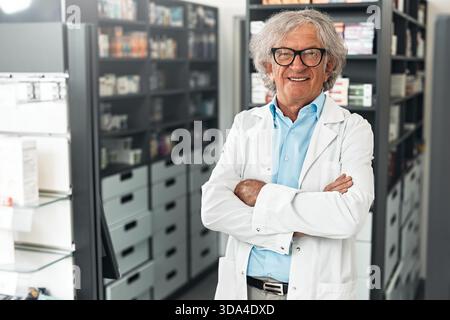 Ritratto di un farmacista anziano sorridente con un cappotto bianco, in piedi dietro il bancone della farmacia. Scaffali con prodotti medicali sullo sfondo. C Foto Stock