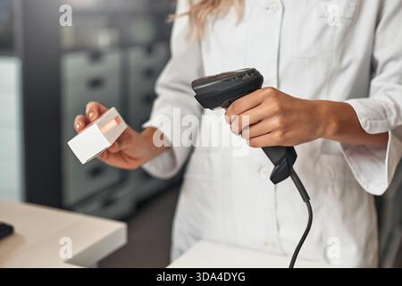 Primo piano del farmacista che esegue la scansione di una cassetta dei medicinali al banco cassa con scanner di codici a barre. Tecnologia sanitaria e concetto del flusso di lavoro della farmacia retail. Foto Stock