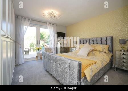 Camera da letto con letto in velluto grigio, coperte gialle e vista del giardino attraverso porte di vetro in una camera soleggiata Foto Stock