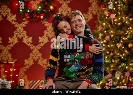 Coppia felice che si avvarrà dell'abbinamento di brutti maglioni natalizi con decorazioni natalizie e luci scintillanti Foto Stock