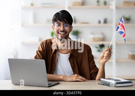 Il giovane sorride mentre tiene in mano una bandiera britannica davanti a un laptop in uno spazio di lavoro luminoso e moderno Foto Stock
