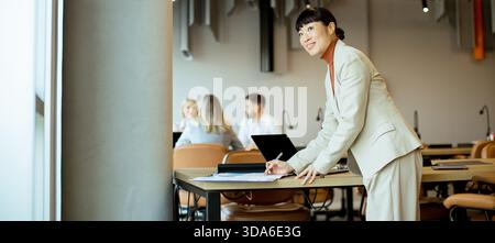 In un ufficio vivace, una donna con un abito scrive a un tavolo mentre i colleghi si impegnano in una conversazione. L'atmosfera è produttiva e concentrata. Foto Stock