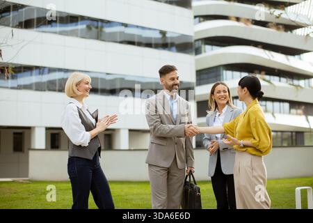 Un gruppo di quattro professionisti si impegna in una stretta di mano amichevole fuori da un elegante edificio di uffici, godendosi un pomeriggio di sole. Foto Stock