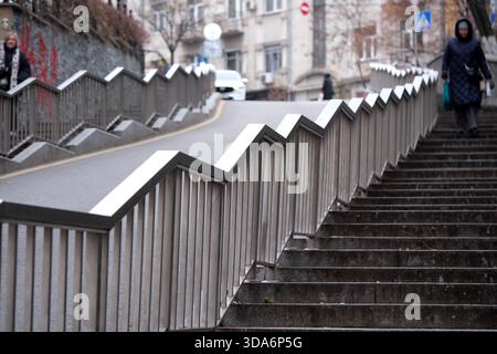 Le persone navigano sui corrimano a zigzag sulle scale, salendo su una tranquilla strada urbana. Kiev, Ucraina. 8 dicembre 2025. Foto Stock