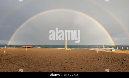 Arcobaleno sopra una spiaggia vuota della Costa Blanca con rete da pallavolo e molo, sabbia bagnata in primo piano dopo la pioggia sotto un cielo grigio nuvoloso Foto Stock
