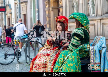 Danzica, Polonia: 2 giugno 2024. Due giovani donne che indossano abiti vivaci e ornati e copricapi decorativi si siedono su una panchina accanto alle biciclette su un vivace cit Foto Stock