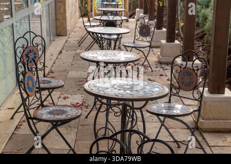 Fila di sedie in ferro battuto e tavoli rotondi a mosaico su uno stretto patio all'esterno di un piccolo caffè, con lastricatrici e foglie cadute, calda atmosfera di luce del giorno, posti a sedere vuoti invitanti pasti all'aperto Foto Stock