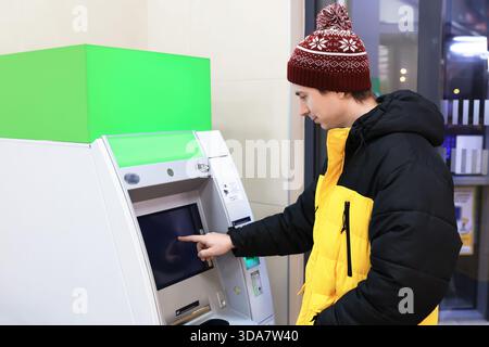 Uomo in abiti invernali che usa bancomat con schermo verde mockup. Persona che ritira denaro al terminale della banca. Foto di alta qualità Foto Stock