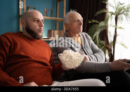 uomo maturo che guarda il campionato con suo figlio adulto che mangia popcorn a casa Foto Stock