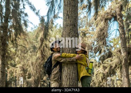 Un uomo e una donna abbracciano un albero in una foresta. L'uomo indossa uno zaino e la donna indossa una giacca gialla Foto Stock