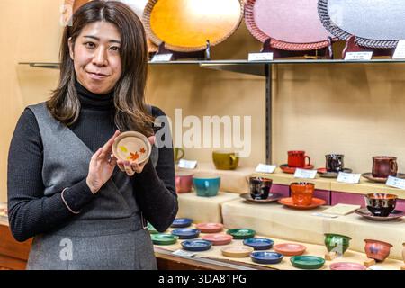 Una giovane donna tiene in mano una piccola ciotola laccata decorata con un motivo di foglie autunnali (momiji) in un negozio di artigianato; sullo sfondo c'è un variopinto assortimento di piccole ciotole e tazze. Shiojiri, Prefettura di Nagano, Giappone Foto Stock
