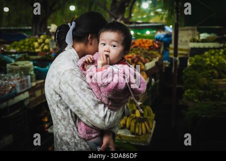 Paro, Bhutan - 17 settembre 2025: Vista di una madre cullando dolcemente il suo bambino nel vivace traffico di un mercato pieno di frutta e verdura colorate Foto Stock