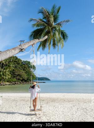 Una splendida spiaggia sull'isola di Koh Kood mostra una donna che si gode un'altalena sotto un'alta palma. Le calme acque blu e il cielo limpido creano una perfetta atmosfera da paradiso tropicale in Thailandia || modello rilasciato Foto Stock