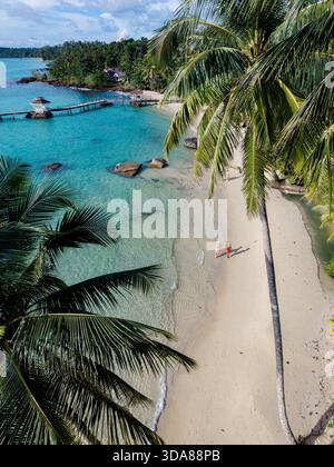 Splendida vista aerea di due persone che camminano lungo la spiaggia sabbiosa dell'isola di Koh Kood in Thailandia. Lussureggianti palme incorniciano la scena, con serene acque turchesi che invitano al relax sotto il sole. Foto Stock