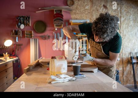 Carpentiere uomo che si prepara a lavorare su nuovi mobili, misura con precisione la quantità richiesta di resina epossidica su bilance elettroniche al tavolo, versare liquido nel bicchiere di carta dal contenitore, processo di officina. || modello rilasciato Foto Stock