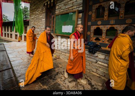 Paro, Bhutan - 17 settembre 2025: Veduta dei monaci con le vivaci vesti arancioni che camminano lungo il sentiero in pietra del monastero di Sangchekhor, la tavola verde è appesa Foto Stock