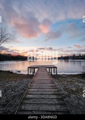 Molo in legno sul lago (vista grandangolare). Scenario invernale all'alba con ghiaccio sull'acqua, alberi e cielo che si riflettono sulla superficie del lago simile a uno specchio. Foto Stock