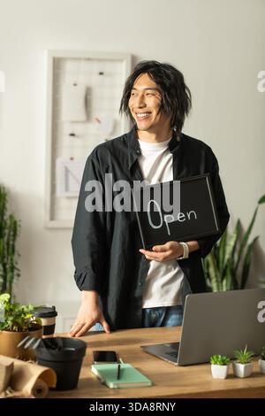 Il giovane fiorista asiatico sta al banco, tenendo un cartello aperto e irradiando felicità. Foto Stock