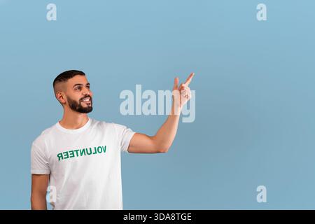 Il giovane uomo che indossa la camicia volontaria sorride e punta verso il cielo in un ambiente luminoso e allegro Foto Stock