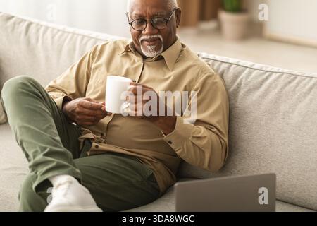 Uomo maturo che si gusta il caffè mentre lavora su un notebook a casa in un luminoso salotto Foto Stock