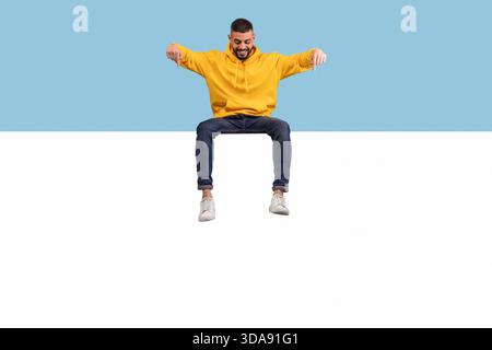 Uomo con cappuccio giallo seduto sul bordo con sfondo blu mentre sorride e guarda sotto Foto Stock