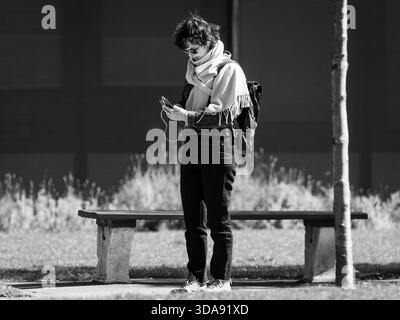 Vienna, Austria - 7 aprile 2020: Una donna si trova in un parco vicino a una panchina e guarda il suo smartphone, catturato in bianco e nero Foto Stock