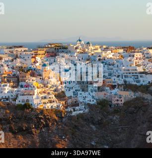 Vista aerea degli iconici edifici bianchi e delle chiese di Oia dalla cupola blu scendono lungo le scogliere verso il Mar Egeo, inondate di calda luce dorata. Foto Stock