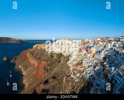 Veduta aerea degli edifici imbiancati che scendono lungo la scogliera verso il mare azzurro, le chiese dalla cupola blu che si stagliano contro il cielo luminoso, Foto Stock
