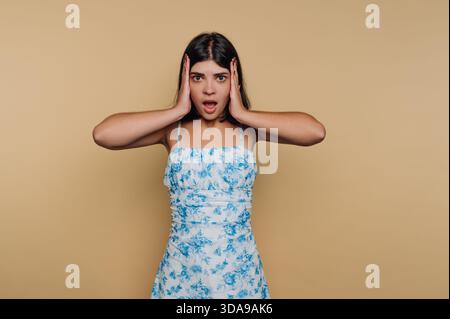 Giovane donna in abito floreale dall'espressione sorpresa, mani sul viso. Foto Stock