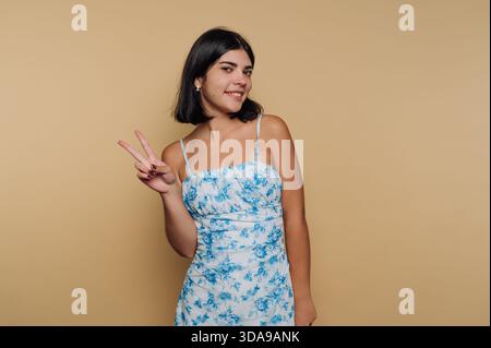 Giovane donna con un abito floreale che mostra un segno di pace su sfondo beige. Foto Stock