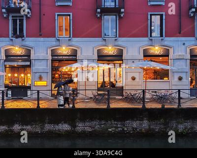 Milano, Italia - 22 maggio 2025: Scena serale piovosa nel quartiere dei Navigli con pedoni sotto ombrelloni che passano davanti al ristorante Mare illuminato Foto Stock