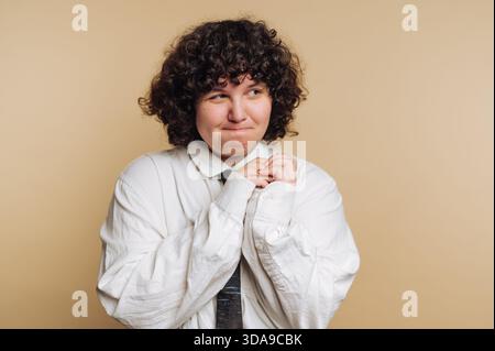 Un giovane adulto dai capelli ricci con un'espressione giocosa si staglia su uno sfondo neutro. Stringono le mani insieme, riflettendo un mix di eccitazione Foto Stock