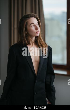Una donna è in piedi vicino a una grande finestra vestita con un blazer nero oversize. La luce naturale illumina la stanza, donandole un tocco di eleganza e sicurezza Foto Stock