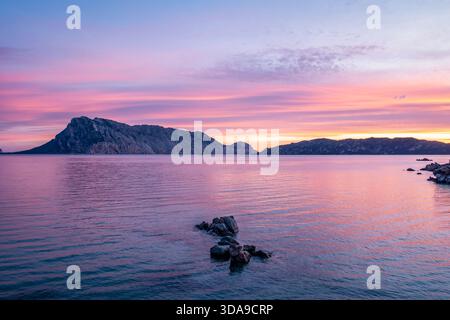 Un tramonto sereno dipinge il cielo in rosa e viola, mentre le acque calme riflettono un lontano orizzonte roccioso dell'isola, Sardegna, Italia Foto Stock