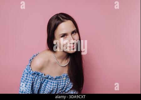 Una donna dai capelli lunghi e lisci sorride dolcemente indossando un top a scacchi blu. Lo sfondo è di colore rosa brillante, creando un pla Foto Stock