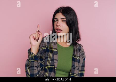 Una giovane donna sta di fronte a una parete rosa, indossando una giacca a quadri sopra una camicia verde. Solleva il dito scherzosamente, mostrando un'esprezione sicura Foto Stock
