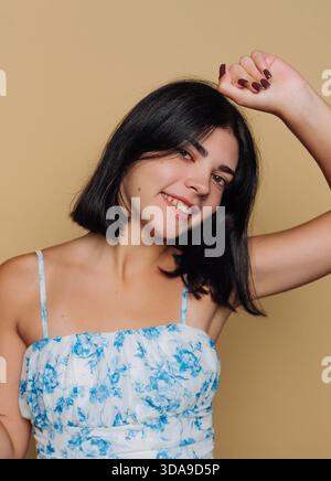 Una giovane donna posa con sicurezza con un sorriso, indossando un abito floreale blu. Migliora il suo comportamento gioioso alzando il braccio, creando un invitante Foto Stock
