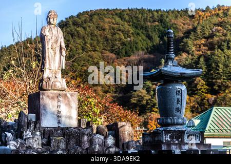 Una statua di Buddha in pietra (Nyorai) e una lanterna in pietra nel cimitero del tempio, con una foresta autunnale di montagna sullo sfondo; l'iscrizione sul plinto identifica la figura come Amida Nyorai, Kiso, prefettura di Nagano, Giappone Foto Stock