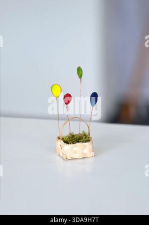 Composizione verticale di un'affascinante scultura in miniatura con quattro palloncini in filo metallico fatti a mano in vivaci colori primari, collocati in un minuscolo cestino intrecciato con Foto Stock
