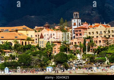 Costa Adeje, Tenerife, Isole Canarie, Spagna, 2023 – gli amanti della spiaggia di fronte a un lussuoso hotel in stile coloniale con montagne sullo sfondo Foto Stock