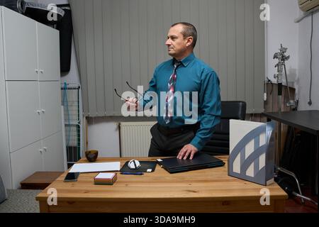 Un uomo d'affari serio e maturo in abbigliamento formale è in piedi vicino alla sua scrivania di legno mentre tiene gli occhiali e guarda lontano Foto Stock