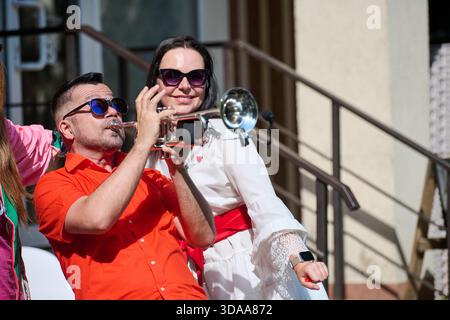 Un uomo adulto che indossa una camicia arancione brillante e occhiali da sole gioca con passione la tromba all'aperto mentre una donna sorridente indossa abiti bianchi e occhiali da sole behi Foto Stock