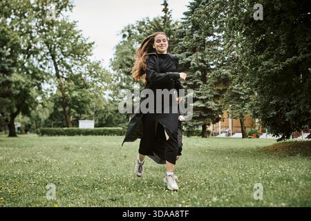 Giovane donna attiva con capelli castani che indossa abbigliamento sportivo nero corre energicamente sull'erba in un parco lussureggiante circondato da alberi. L'atmosfera è vivace e rinfrescante Foto Stock