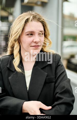 Una giovane donna con i capelli biondi ondulati sta all'aperto e indossa un blazer nero. Si abbina perfettamente alla luce naturale e soffusa del giorno. Blurr Foto Stock