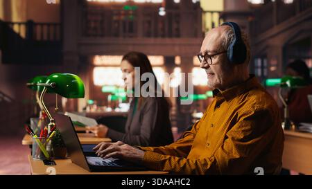 Adulto anziano che disturba le altre persone in biblioteca con il suo rumore musicale, ascoltando musica ad alto volume e cantando alla scrivania. Un uomo anziano che rompe le regole della biblioteca con canzoni solide. Foto Stock